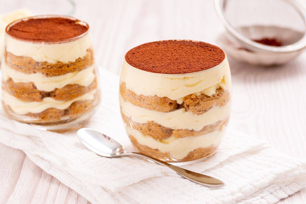 Two glass cups of layered tiramisu dessert with cocoa powder on top sit on a white cloth, accompanied by a metal spoon.