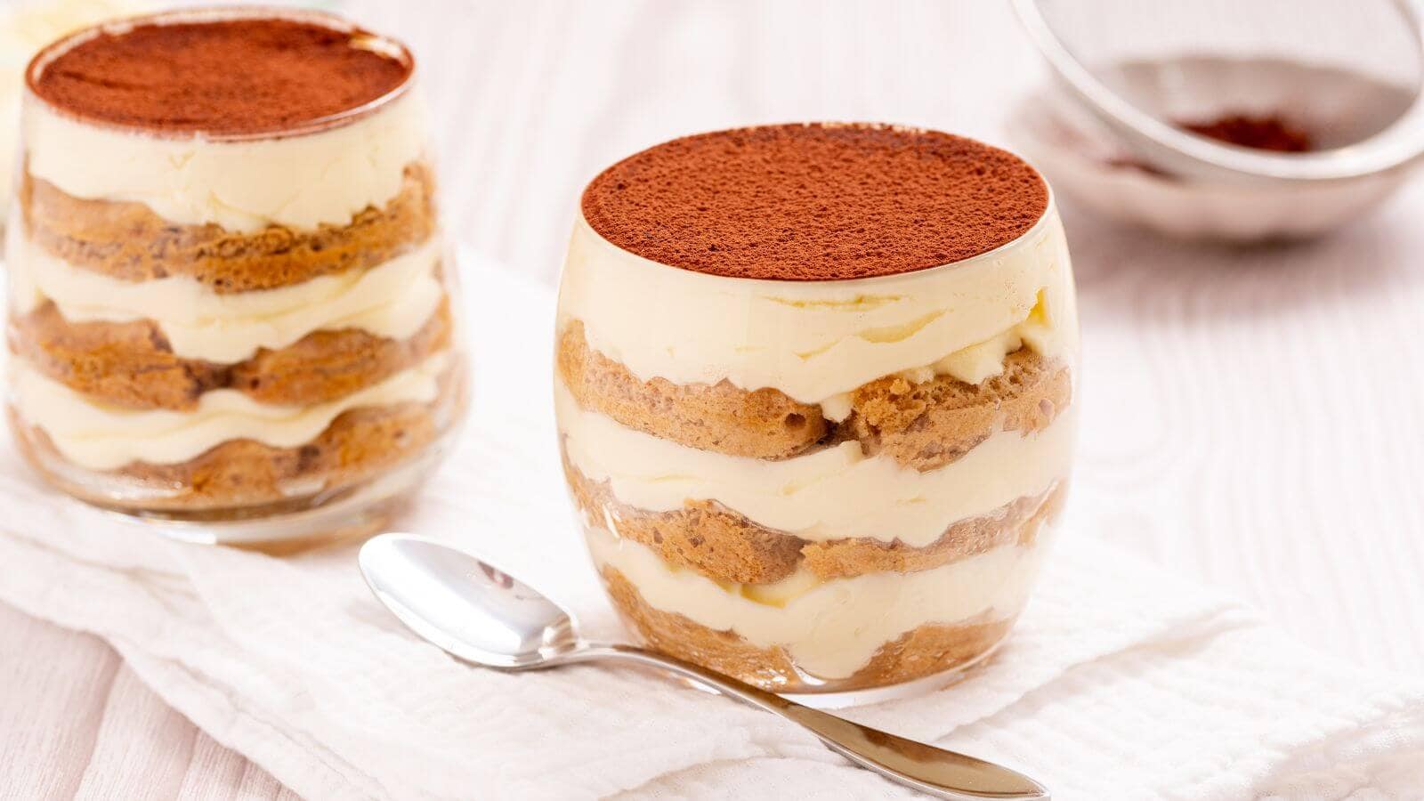 Two glasses of tiramisu dessert with layers of mascarpone cheese and coffee-soaked ladyfingers topped with cocoa powder, placed on a white cloth next to a spoon.