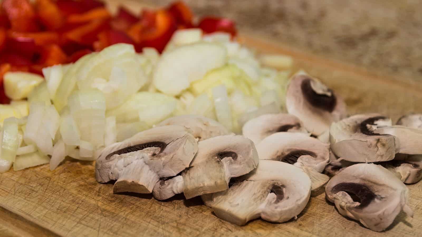 Chopped red bell peppers, onions, and sliced mushrooms on a wooden cutting board.