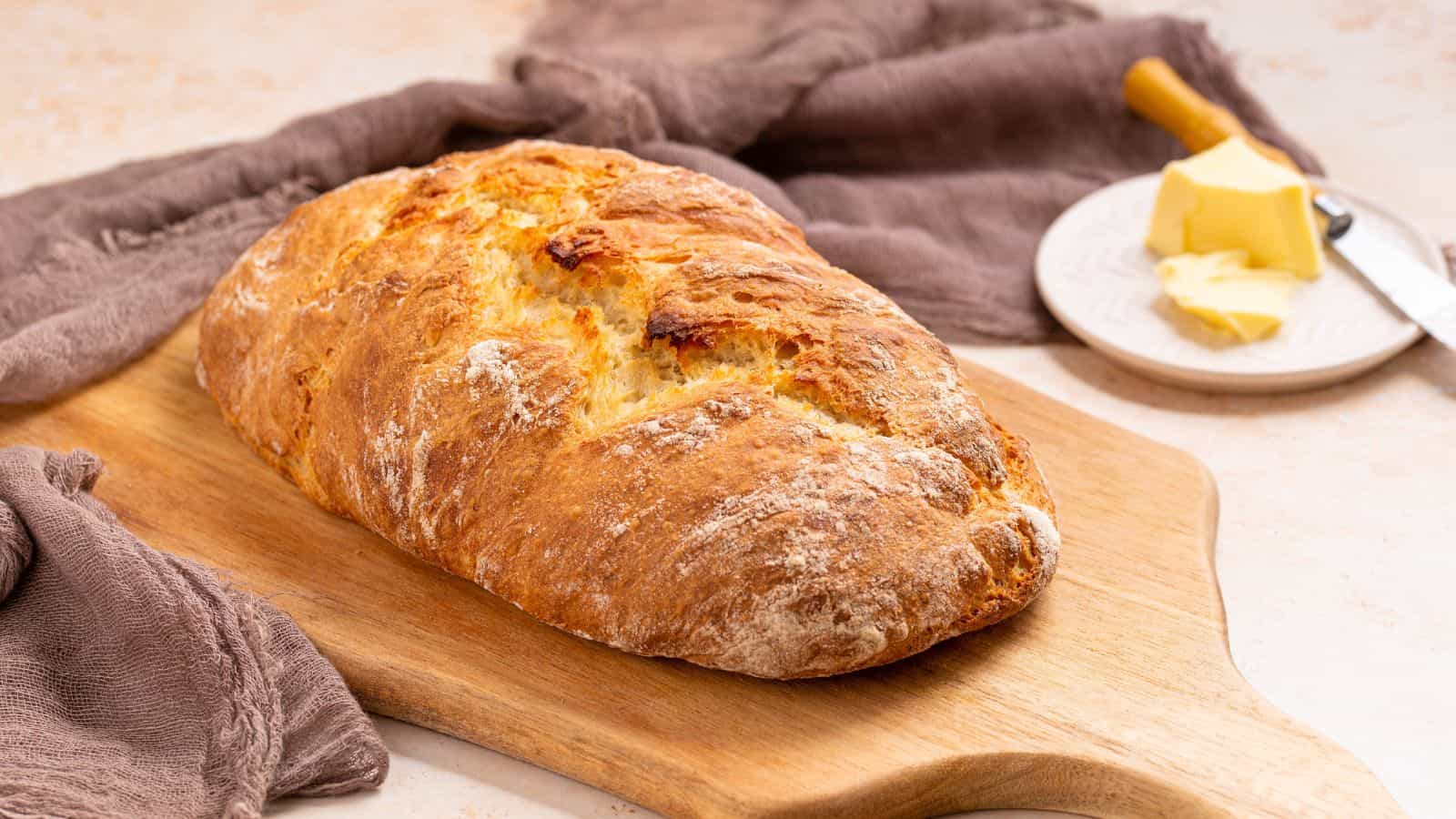 A freshly baked loaf of crusty bread on a wooden cutting board, with a small plate of butter and a butter knife beside it. Brown cloth napkins are placed around the board.