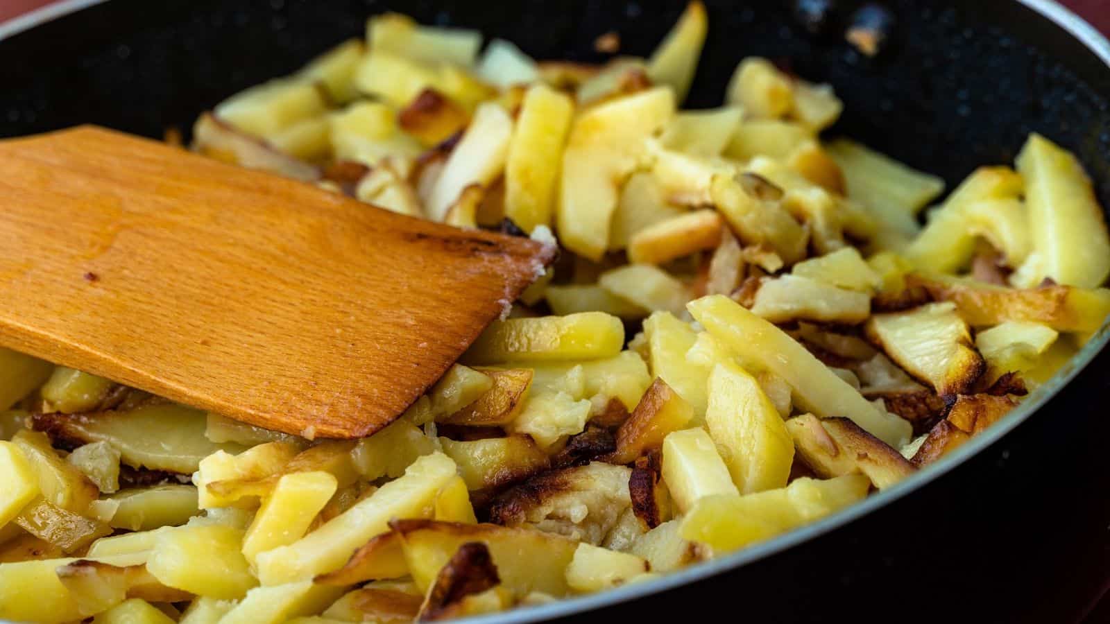 A wooden spatula stirring sliced fried potatoes in a black skillet.
