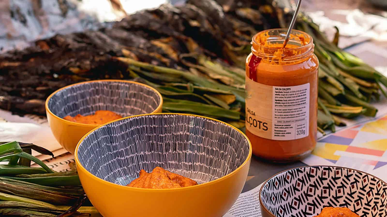Close-up of three bowls containing a red sauce sitting on a colorful tablecloth, with a jar of the same sauce and grilled green vegetables positioned in the background.
