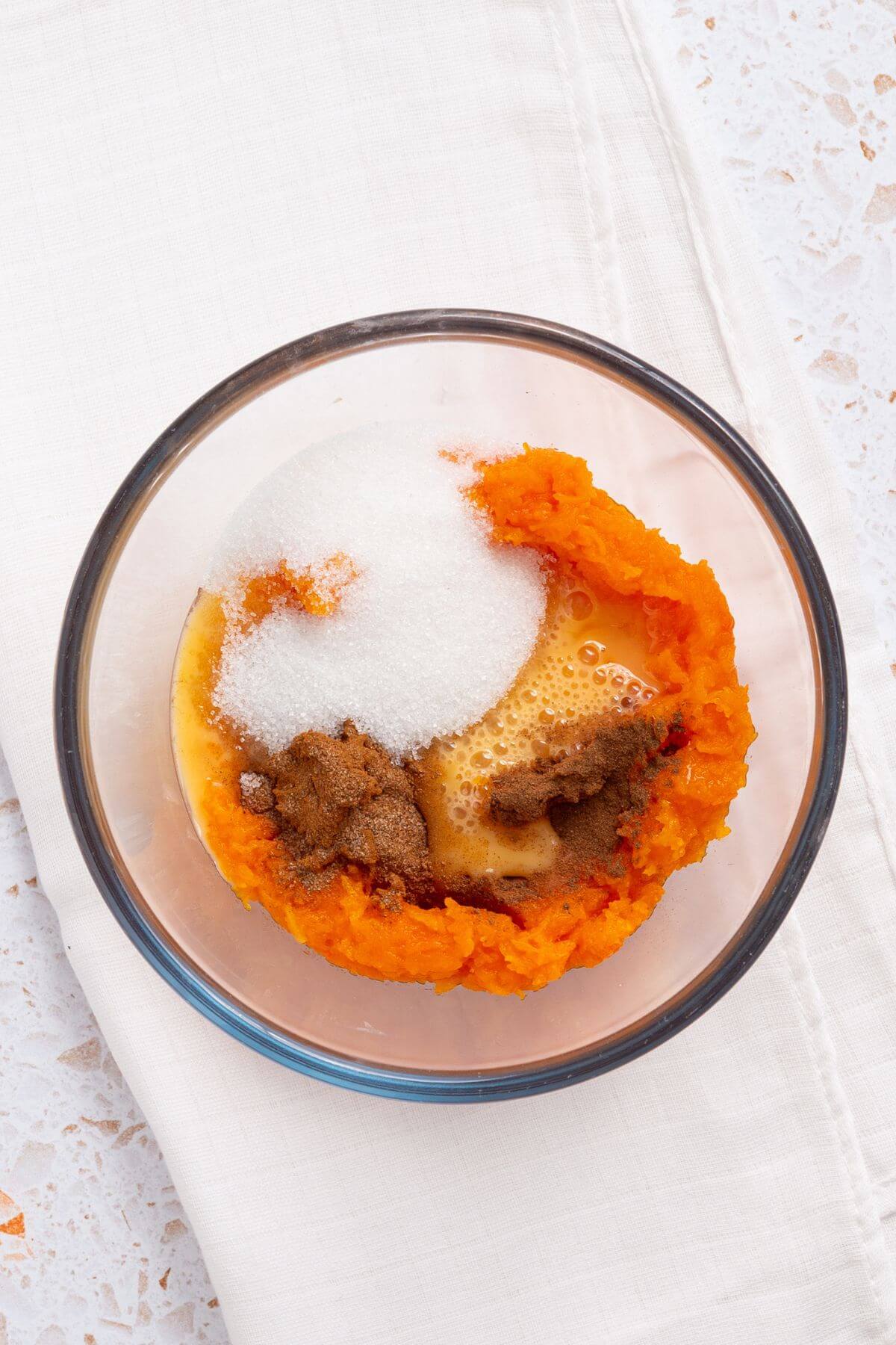 A clear glass bowl containing pumpkin puree, granulated sugar, spices, and beaten eggs sits on a white cloth surface.