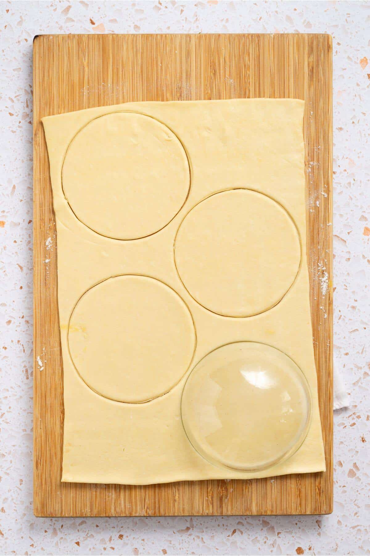 Rectangular sheet of dough on a wooden board with four circle cutouts for pumpkin hand pies. A glass bowl nearby suggests it was used for cutting.