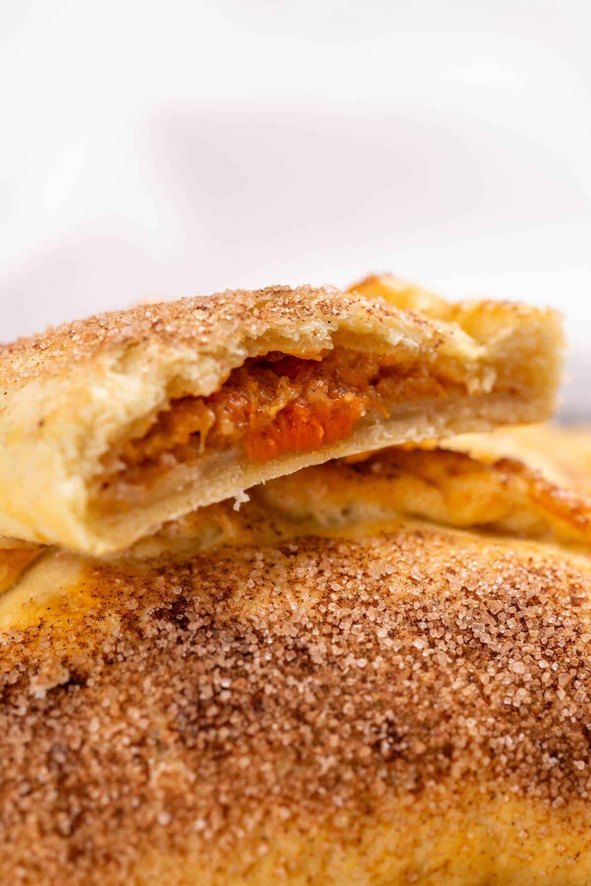 Close-up of a baked pumpkin hand pie with a cinnamon-sugar crust, cut in half to reveal its vibrant orange filling.