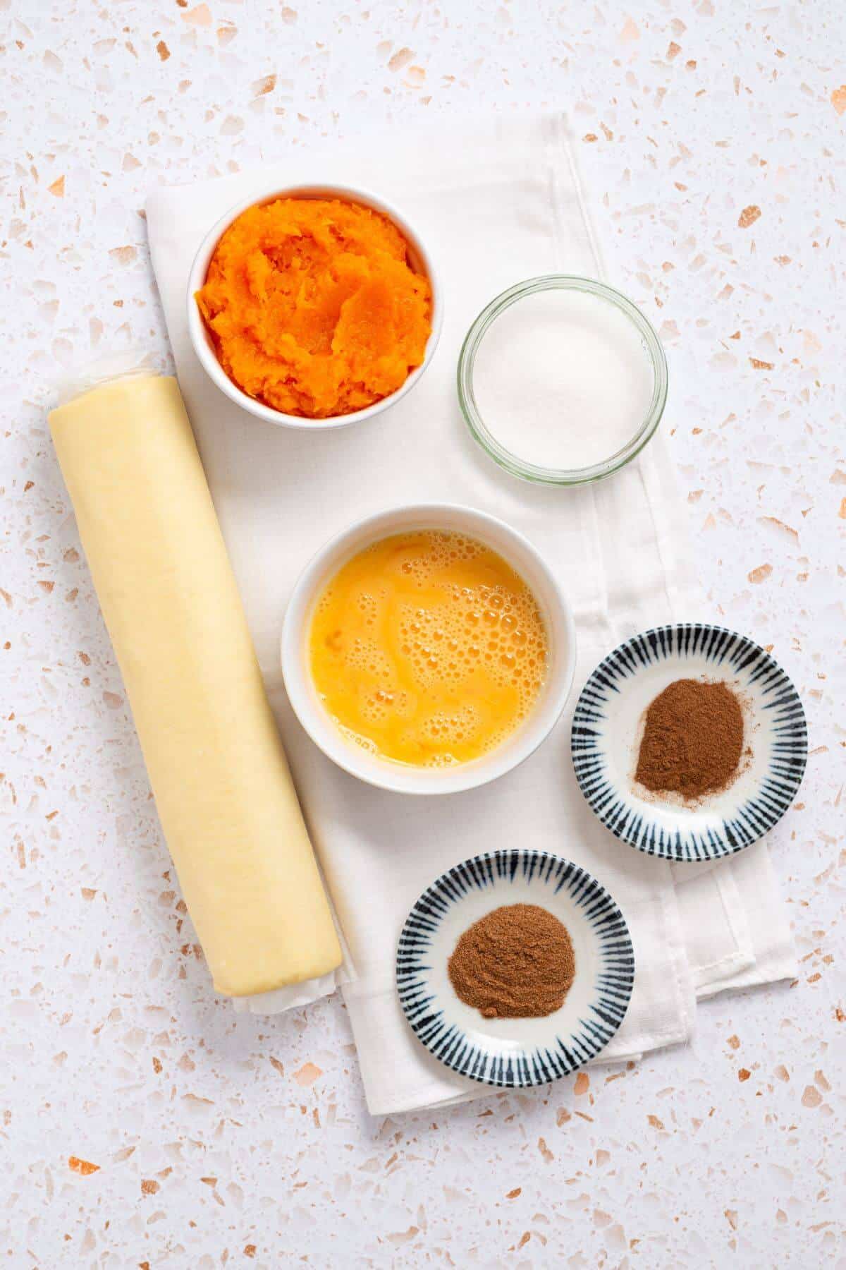 A rolled-out pastry dough next to bowls containing pumpkin puree, beaten eggs, sugar, and small dishes with spices, all arranged on a white cloth over a speckled countertop-perfect for making pumpkin hand pies.
