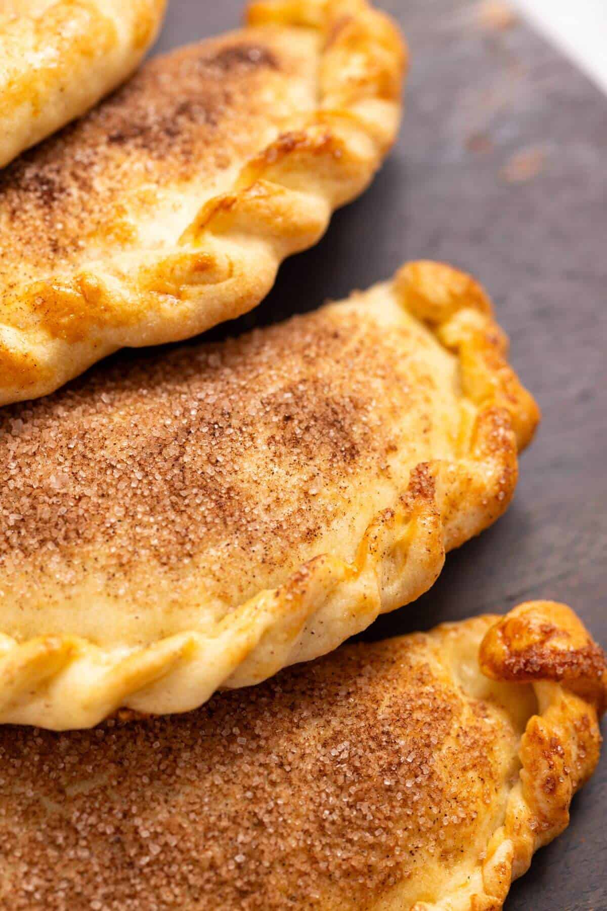Close-up image of three pumpkin hand pies dusted with a cinnamon-sugar mixture, showcasing their golden-brown, flaky crusts and crimped edges.