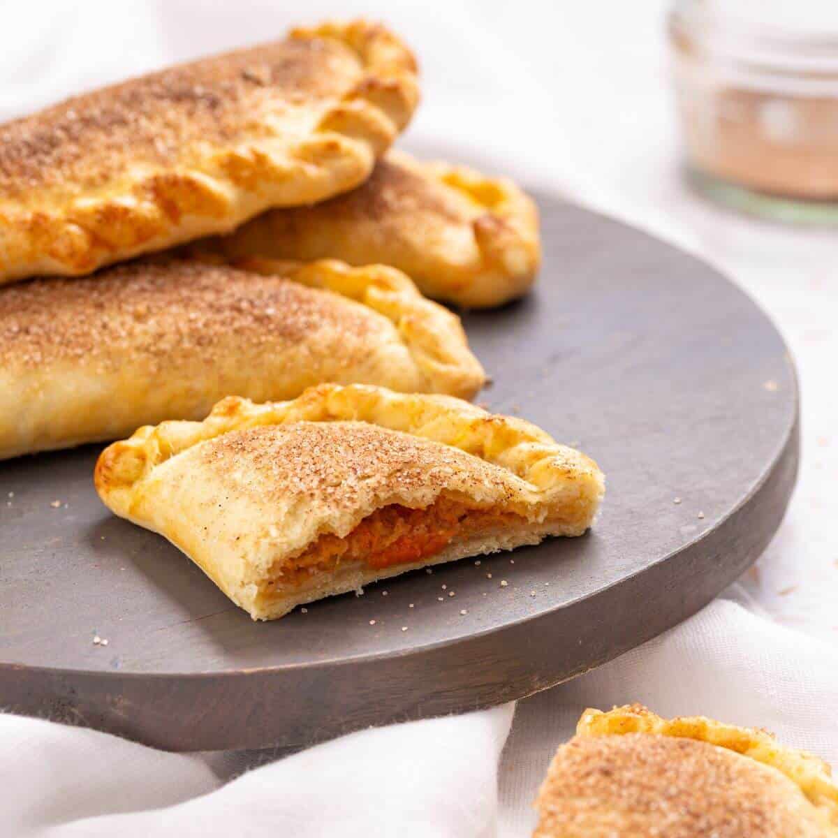 A plate of pumpkin hand pies, with one showing its filling, displayed on a round slate board.