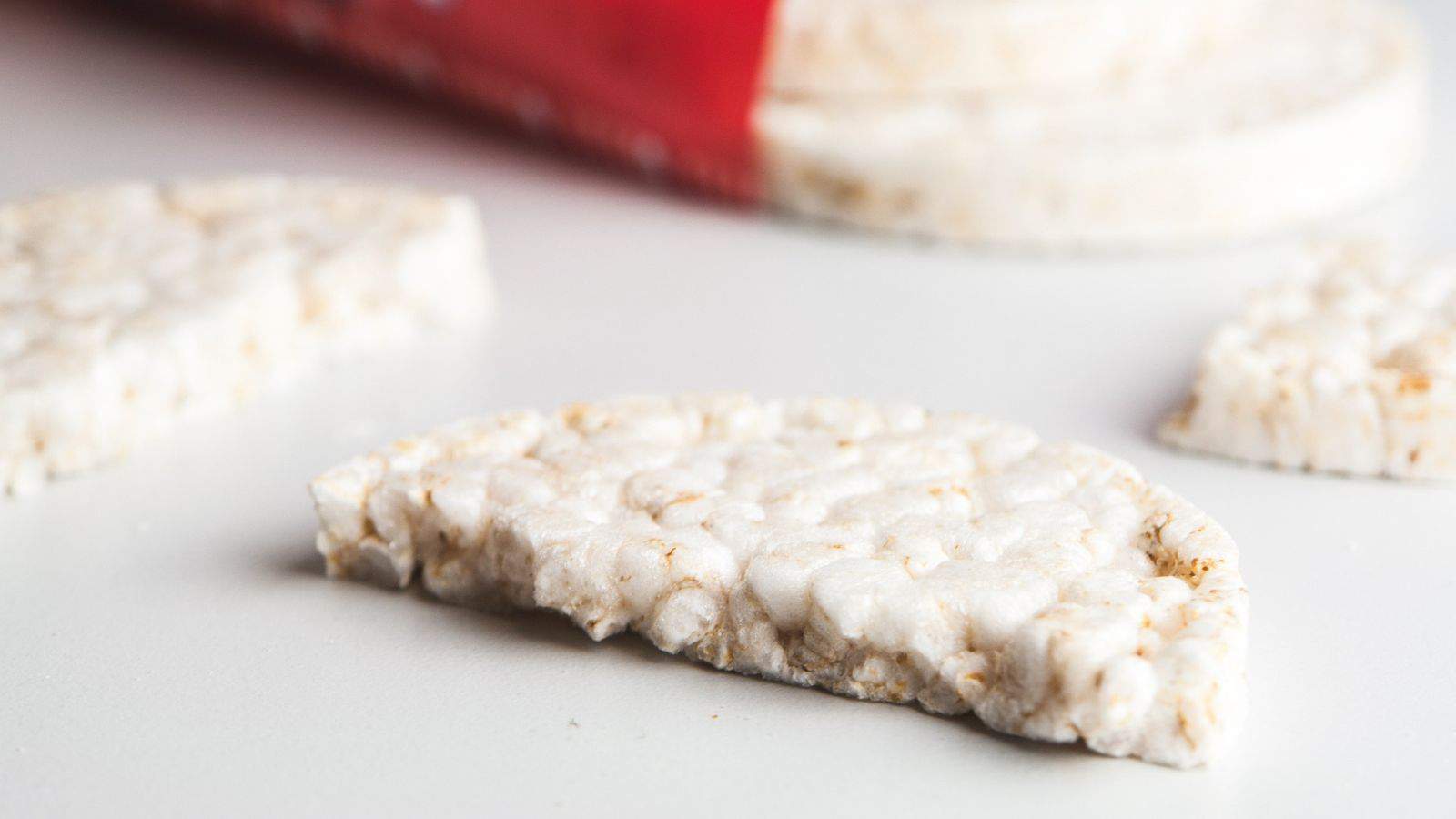 Close-up of a broken rice cake on a white surface, with a package and more rice cakes in the background.