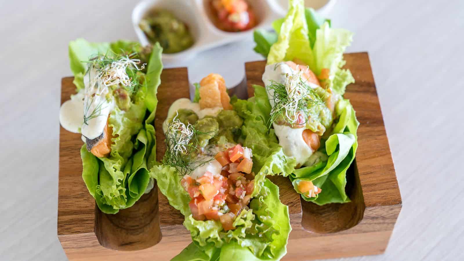 Three lettuce wrap tacos filled with various ingredients including diced tomatoes, sauce, and sprouts, are presented on a wooden tray, with a small dish of additional condiments in the background.