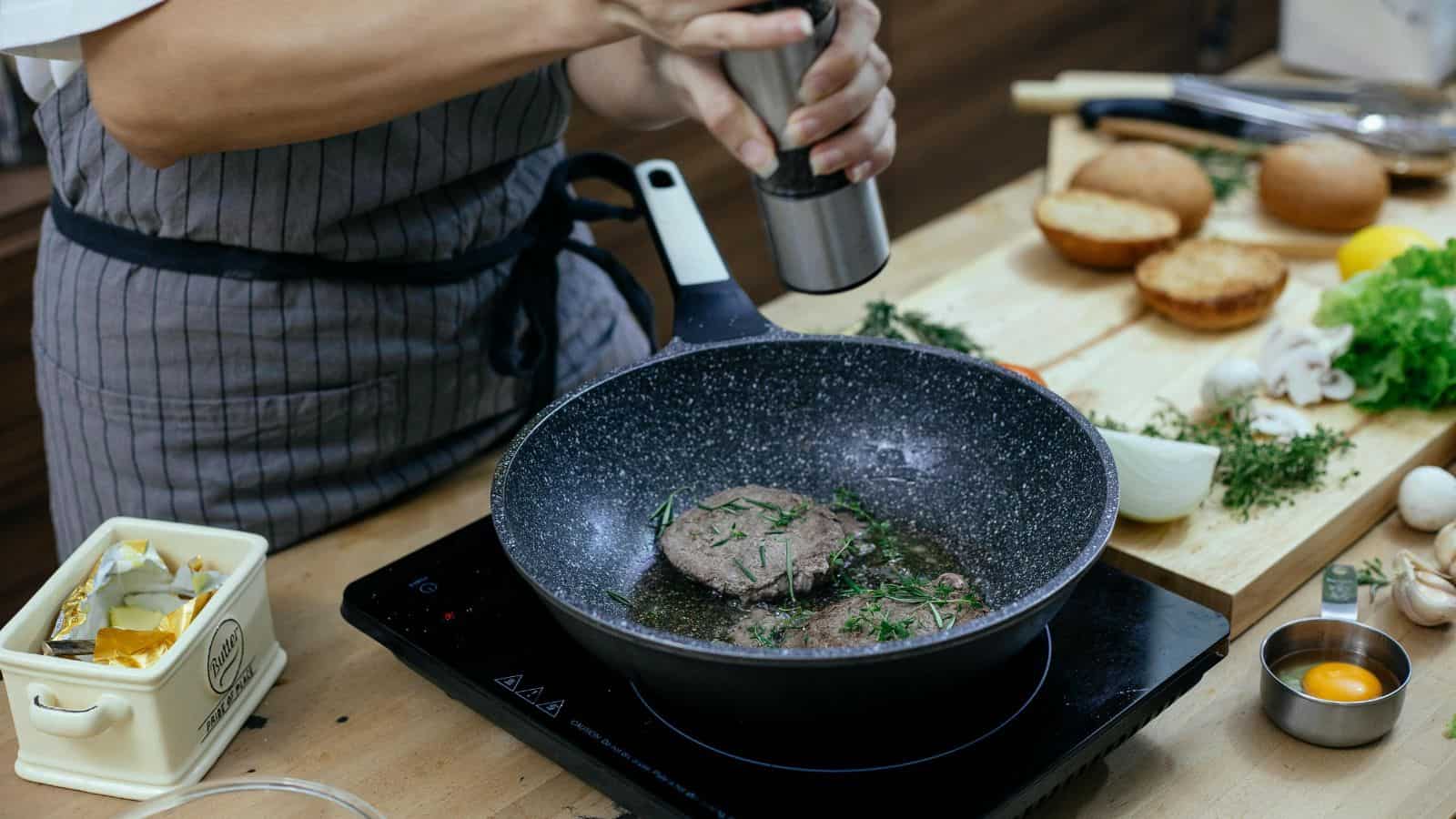 Person seasoning a beef patty in a pan on an induction cooker, surrounded by various ingredients including butter, buns, onions, and lettuce on a wooden kitchen counter.