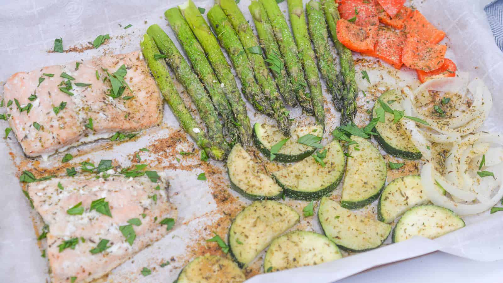 A parchment-lined baking sheet with seasoned salmon, asparagus, bell pepper strips, zucchini slices, and onion rings, all sprinkled with herbs and spices.
