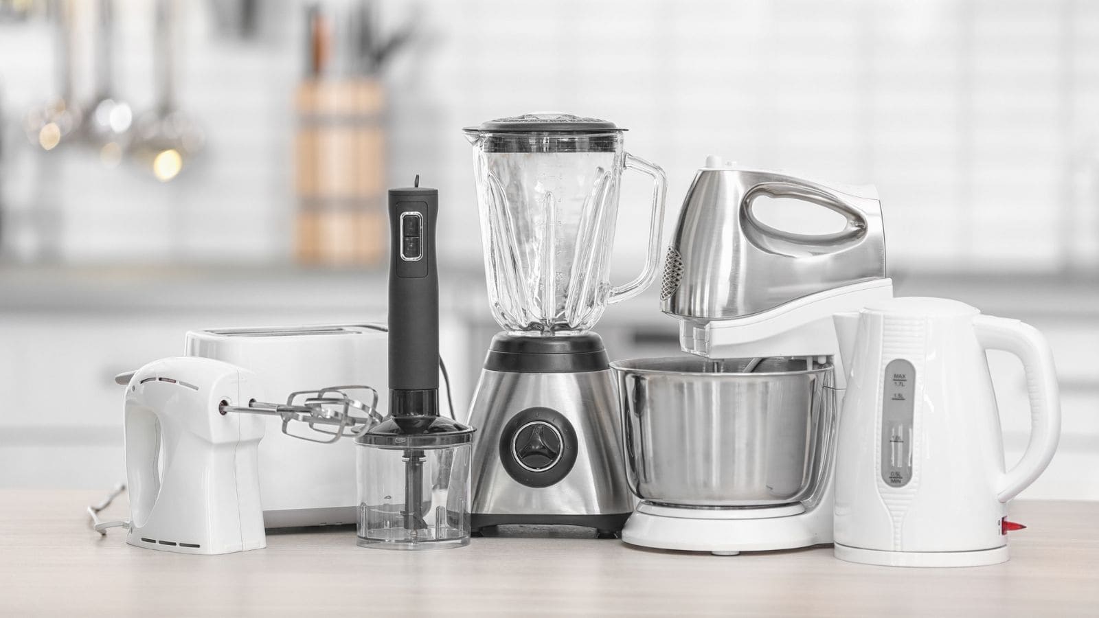 A variety of kitchen appliances, including a blender, kettle, hand mixer, stand mixer, toaster, and hand blender arranged on a kitchen counter, perfect for preparing foods you should always have in your pantry.