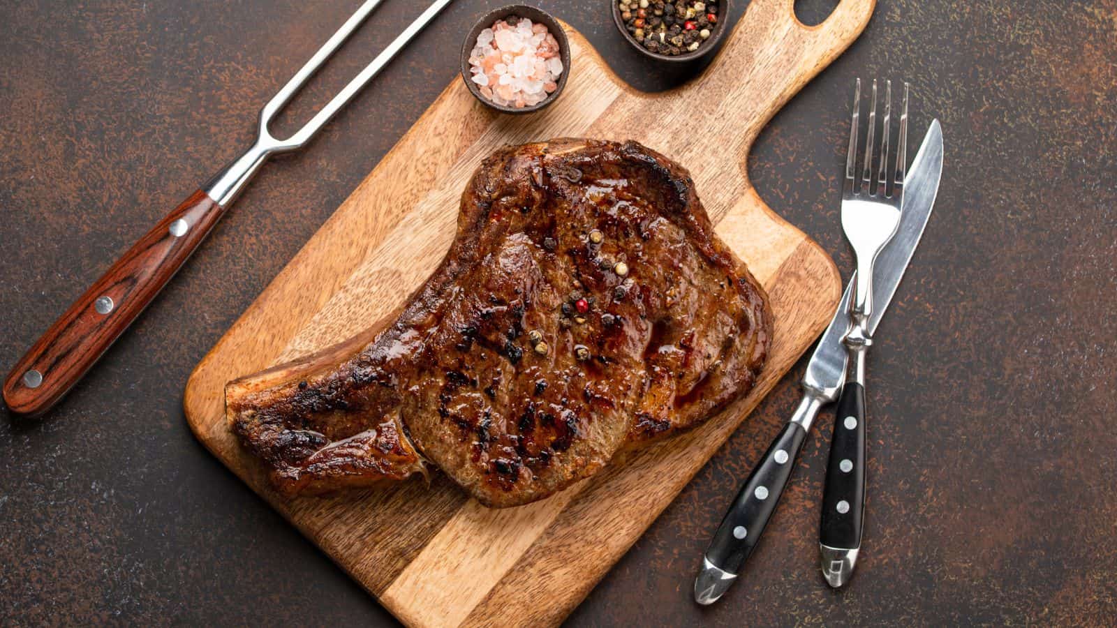 A grilled steak on a wooden cutting board, accompanied by a carving fork, knife, a bowl of salt, and a bowl of mixed peppercorns.