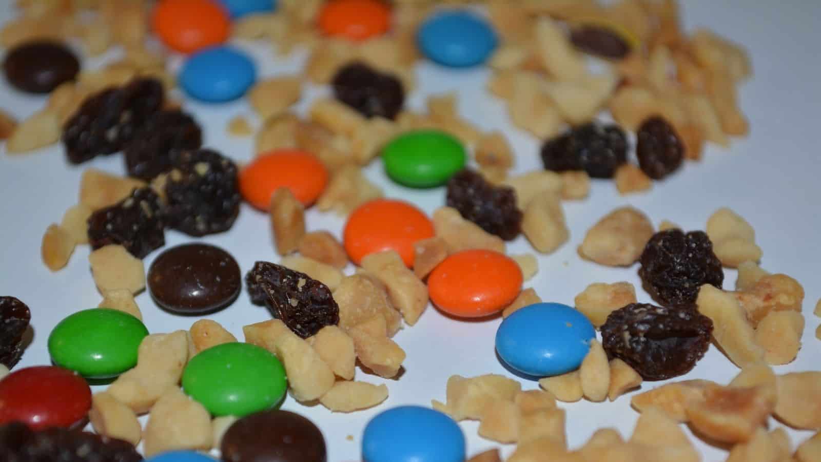 A close-up of a trail mix scattered on a white surface, containing M&M candies in various colors, pieces of peanuts, and raisins.