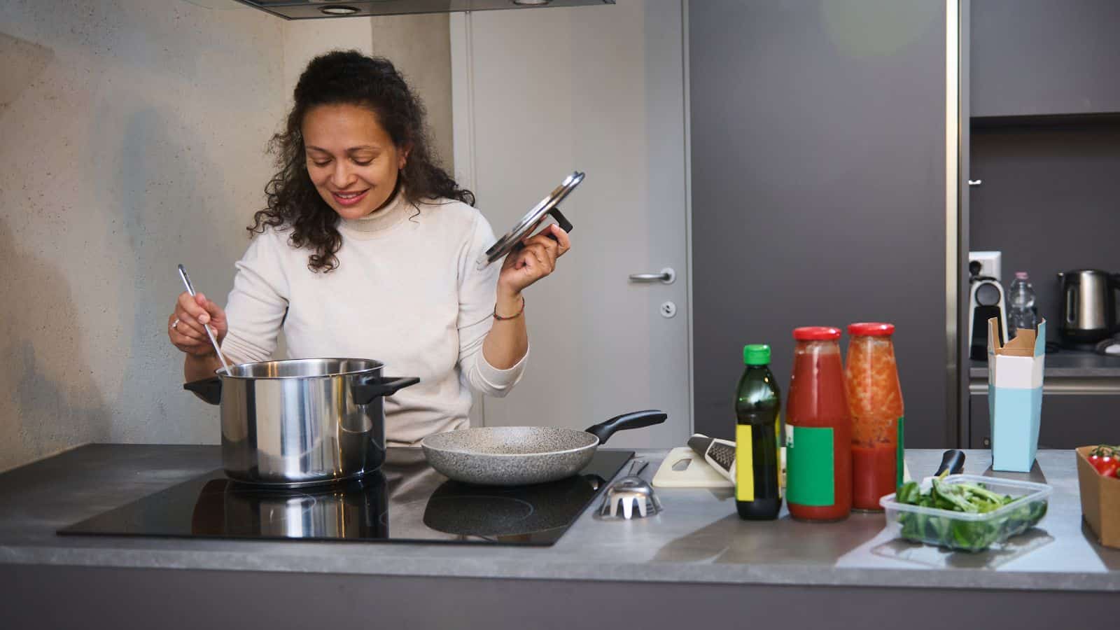 A person smiles while stirring a pot on the stove in a kitchen. Nearby are cooking ingredients including bottled sauces, a frying pan, and fresh greens.