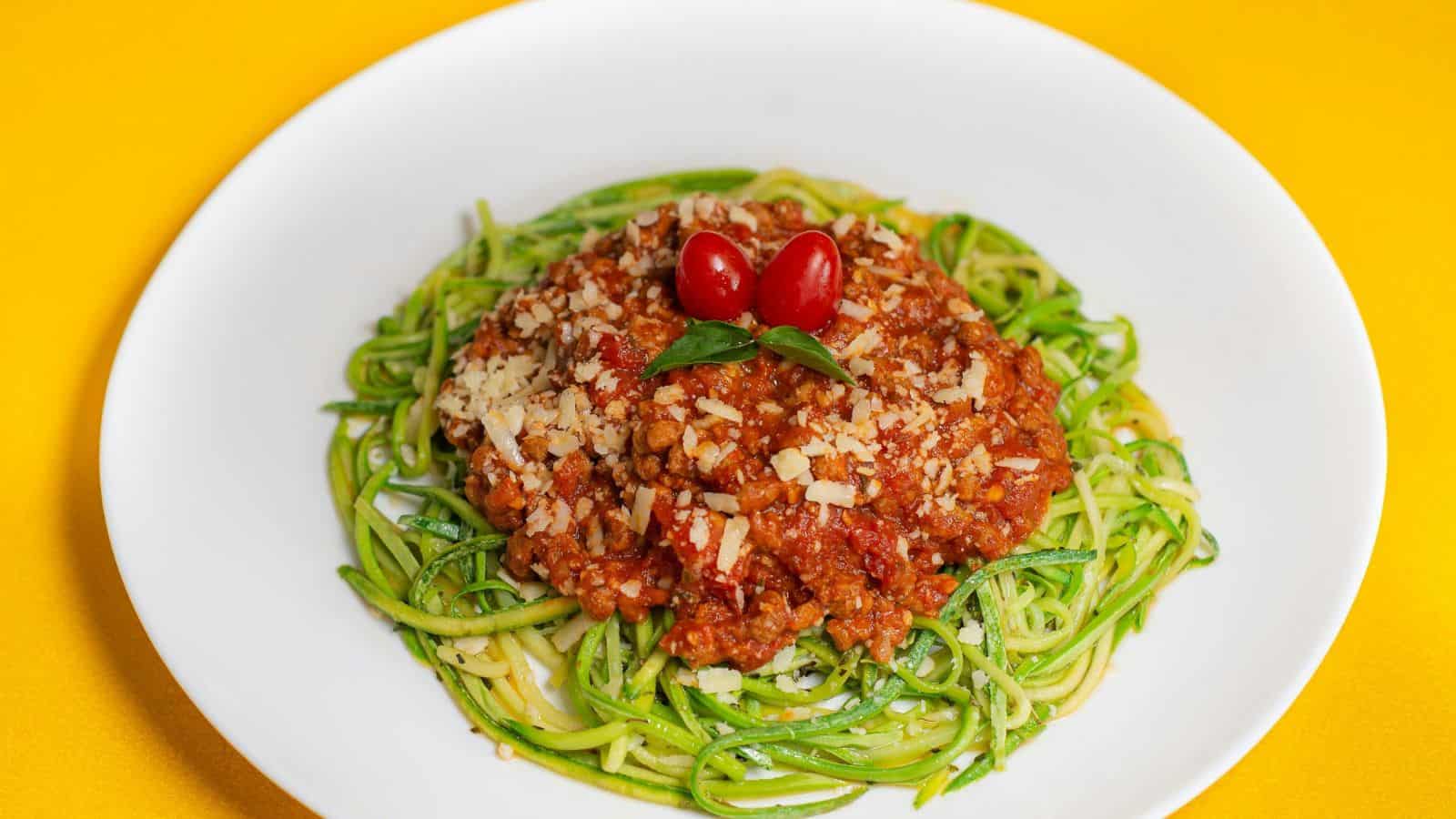 A white plate with zucchini noodles topped with a generous amount of tomato-based meat sauce, garnished with grated cheese, basil leaves, and two cherry tomatoes. Yellow background.
