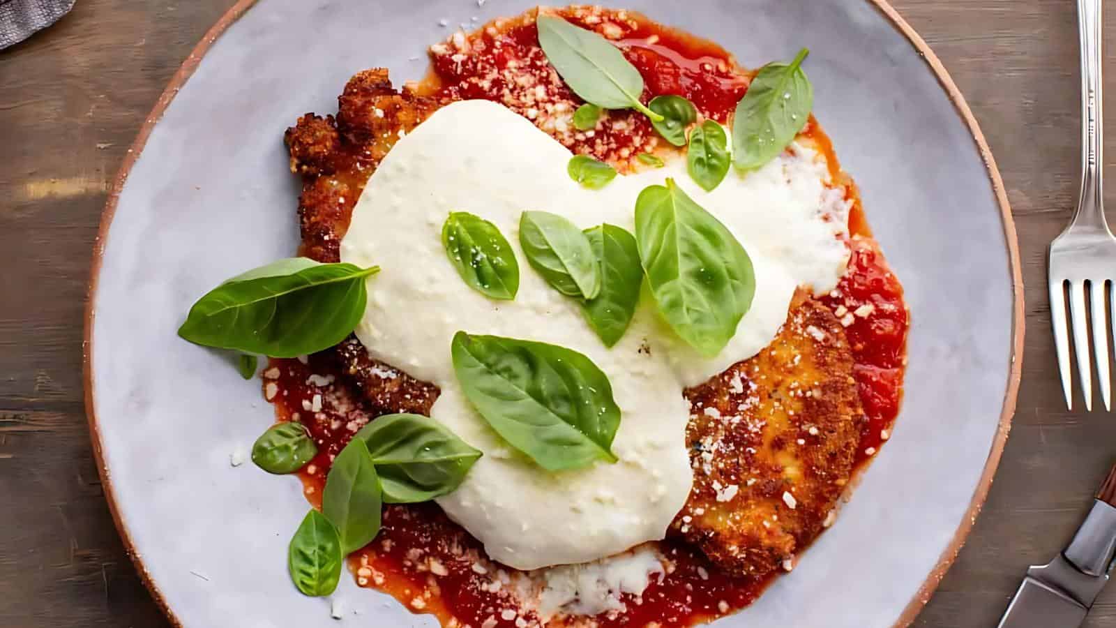 A plate of chicken parmesan topped with melted cheese and fresh basil sits on a layer of tomato sauce. A fork is placed to the right.