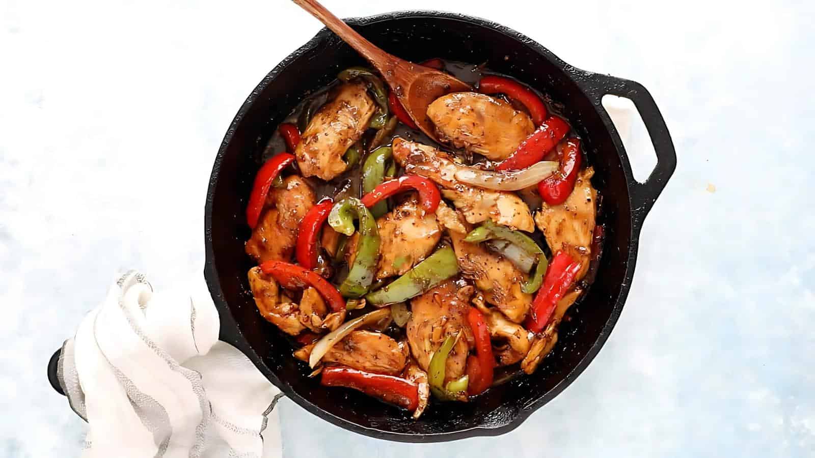 A skillet with cooked chicken pieces, red and green bell peppers, and onions, stirred with a wooden spoon.