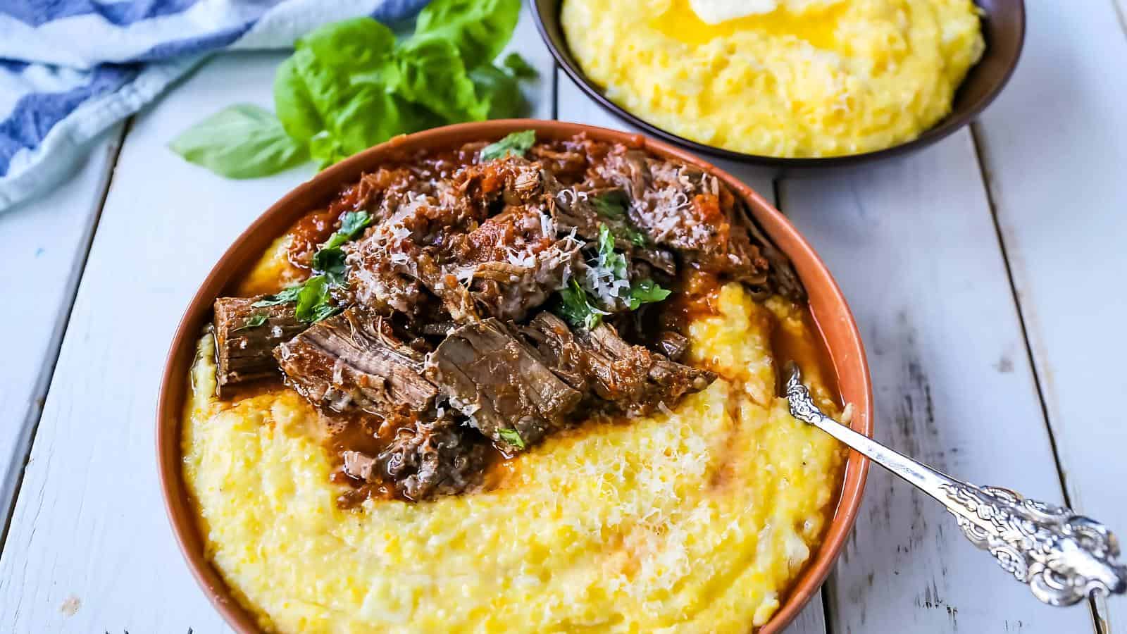 A bowl of cheesy polenta topped with shredded beef and herbs beside another bowl of plain polenta on a white wooden table.