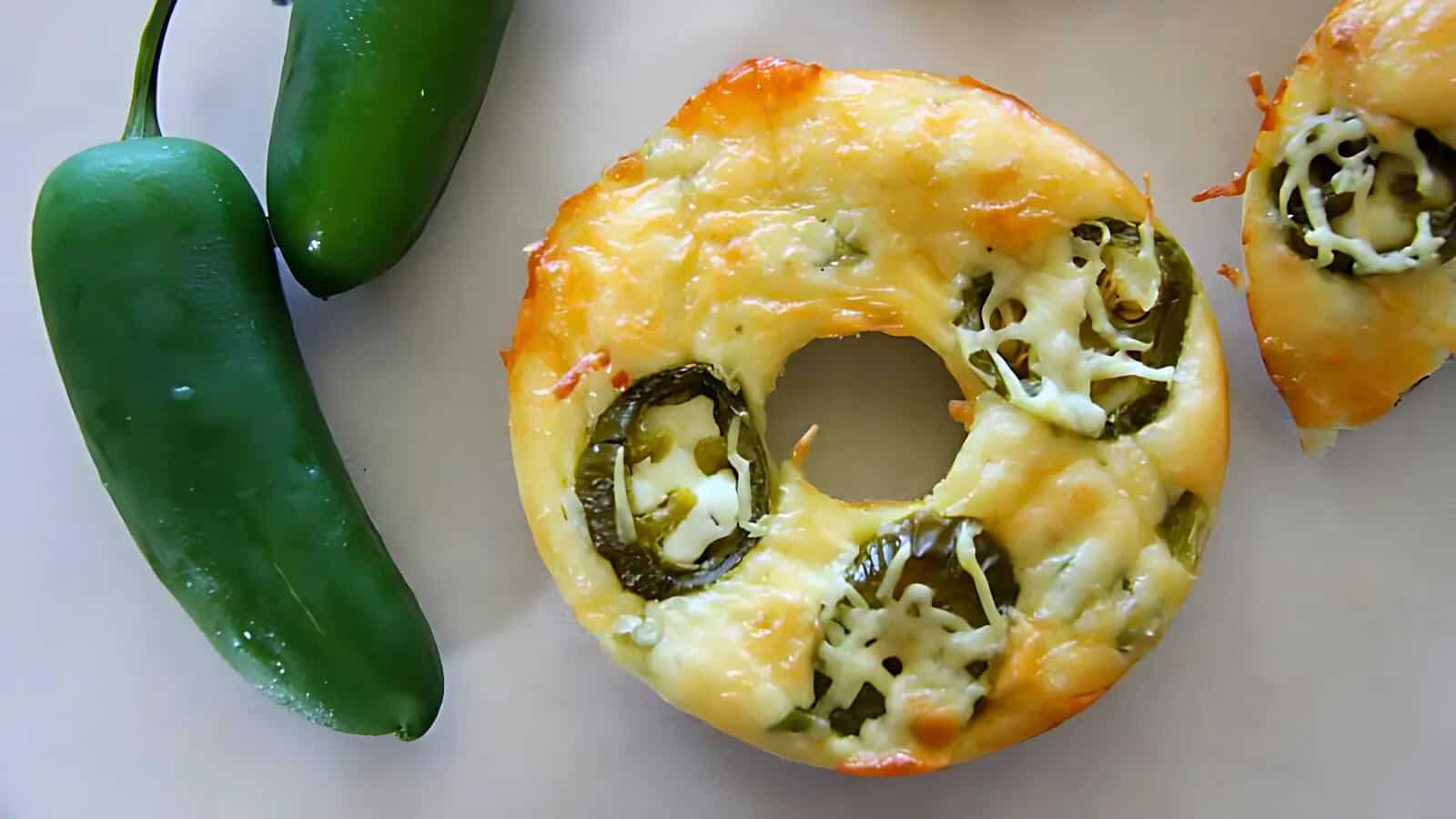 A baked bagel topped with cheese and jalapeño slices, next to two whole jalapeños on a white surface.