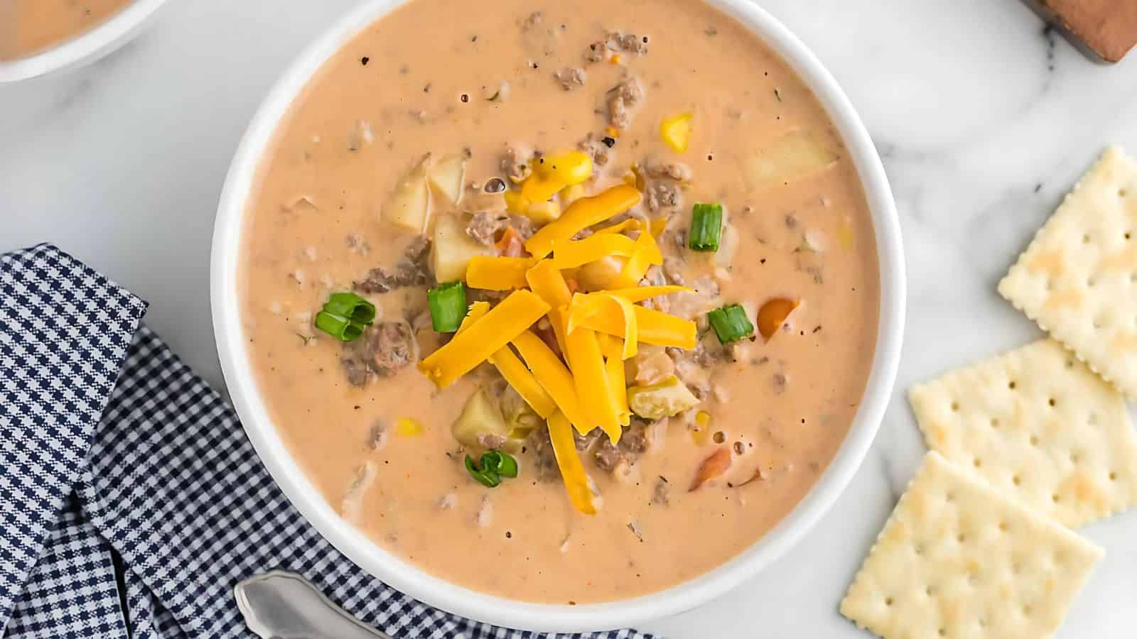 Bowl of cheeseburger soup topped with cheddar cheese and green onions, next to crackers on a checkered napkin.