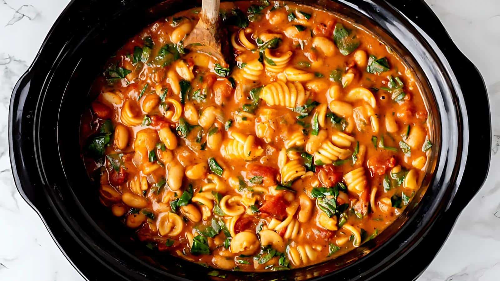 A slow cooker filled with spiral pasta in a creamy tomato sauce, mixed with beans and spinach, and stirred with a wooden spoon.
