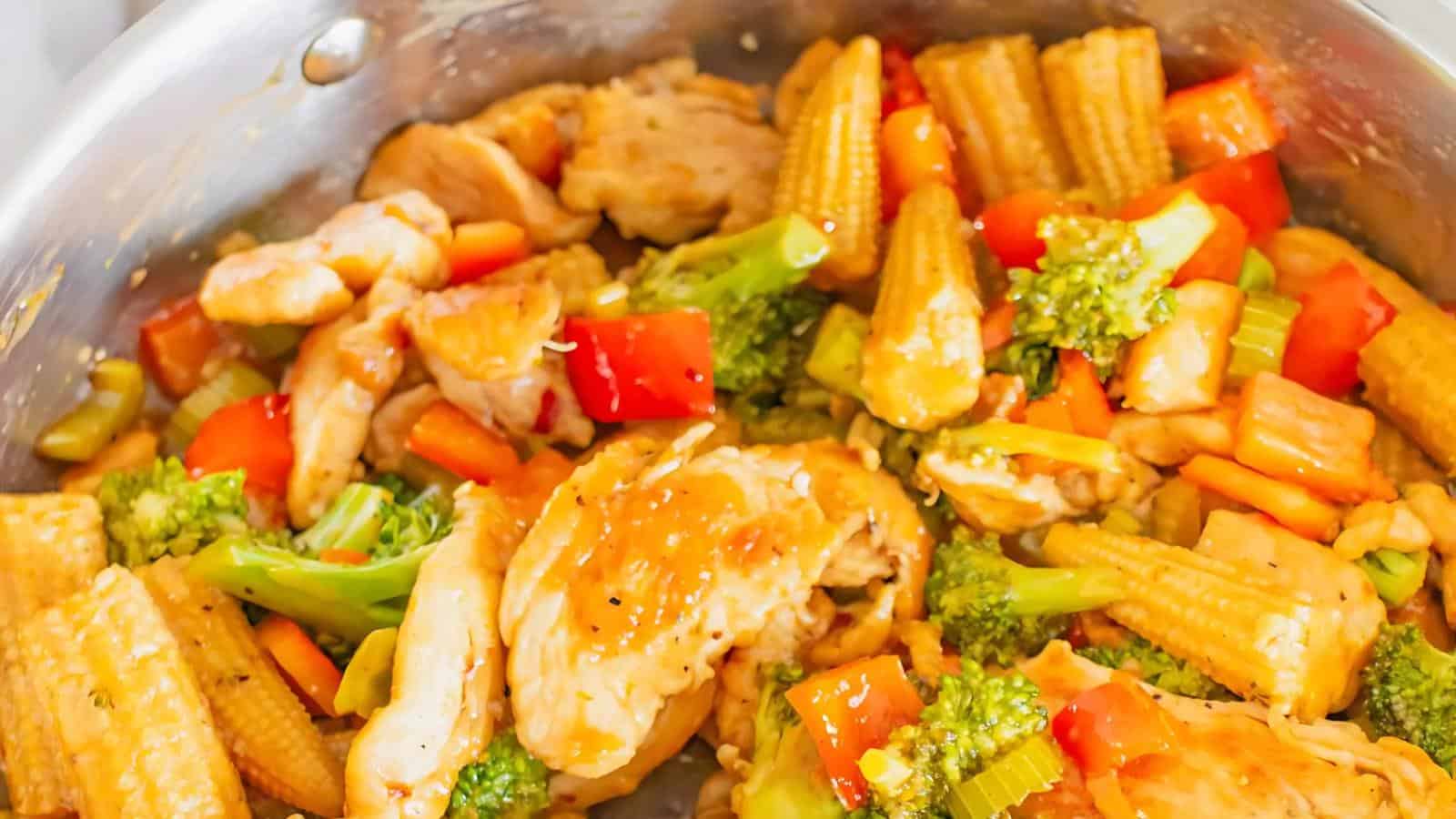 A stir-fry dish with sliced chicken, baby corn, broccoli, red bell peppers, and carrots in a pan.