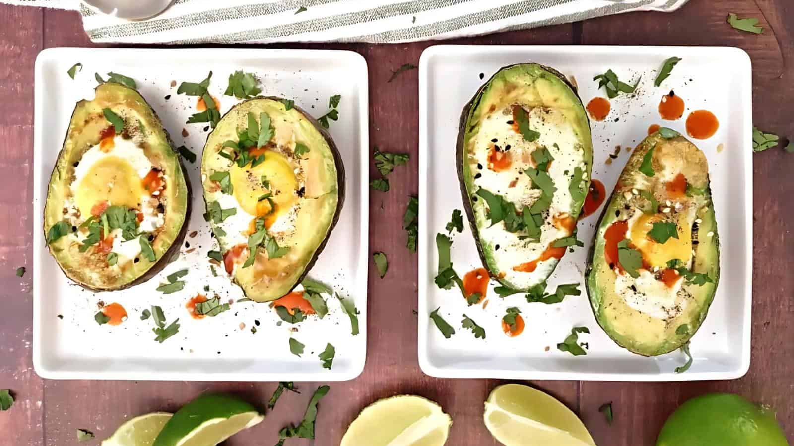 Two square plates each with a baked avocado half, filled with a cooked egg, garnished with cilantro and hot sauce, placed on a wooden surface with lime wedges nearby.
