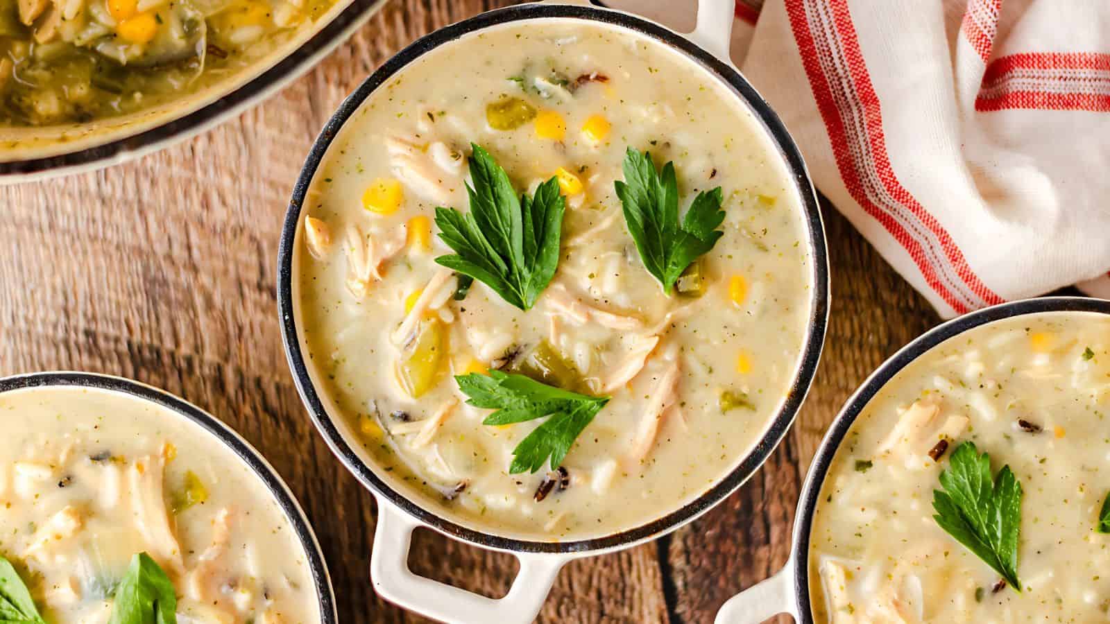 Bowls of creamy soup with chicken, corn, and garnished with parsley on a wooden surface, next to a white and red striped cloth.