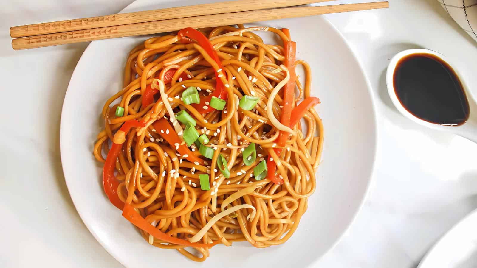 A plate of lo mein with sliced red bell peppers and green onions, garnished with sesame seeds. Chopsticks rest on the plate, and a small dish of soy sauce is beside it.