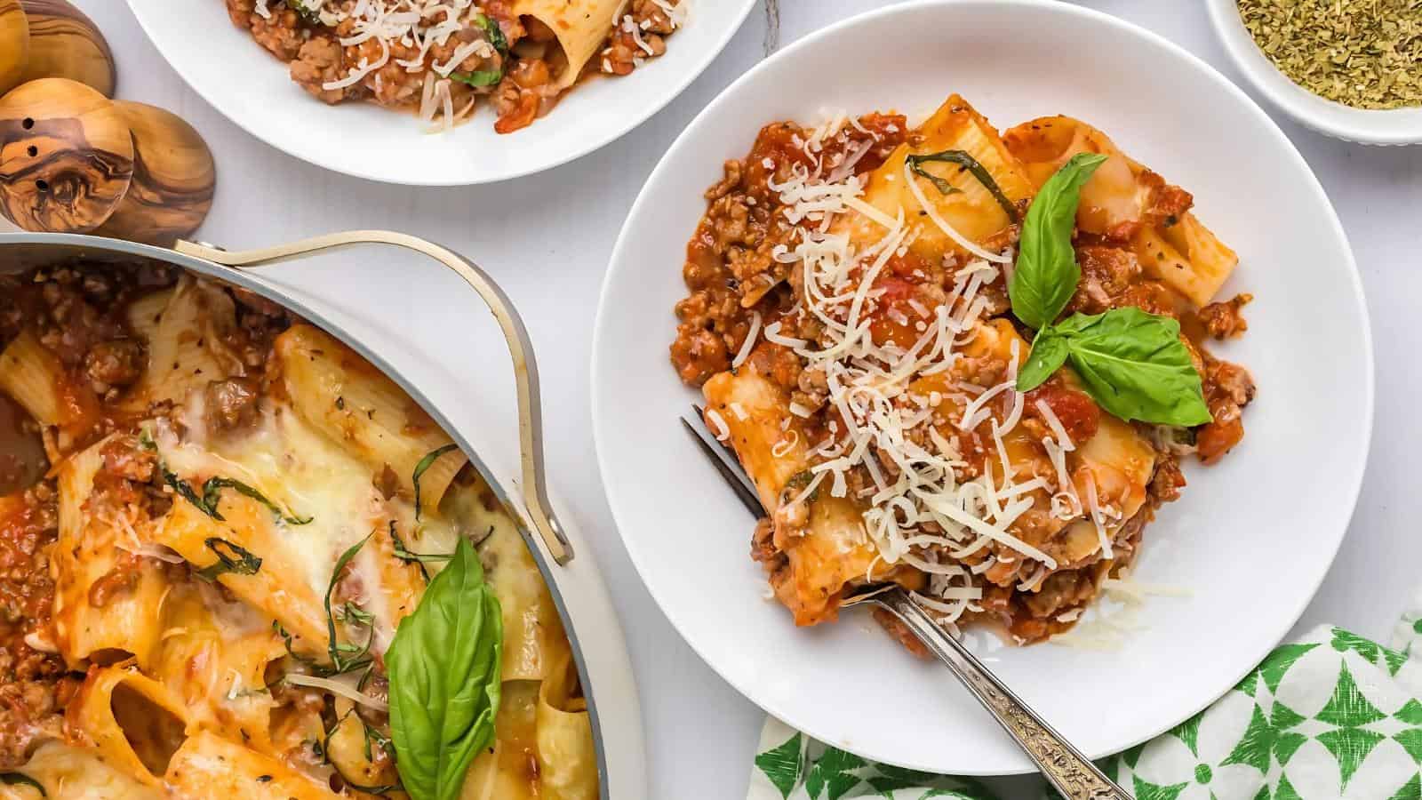 Plates of baked ziti topped with cheese and basil leaves, accompanied by a pan of the same dish and a small bowl with spices.