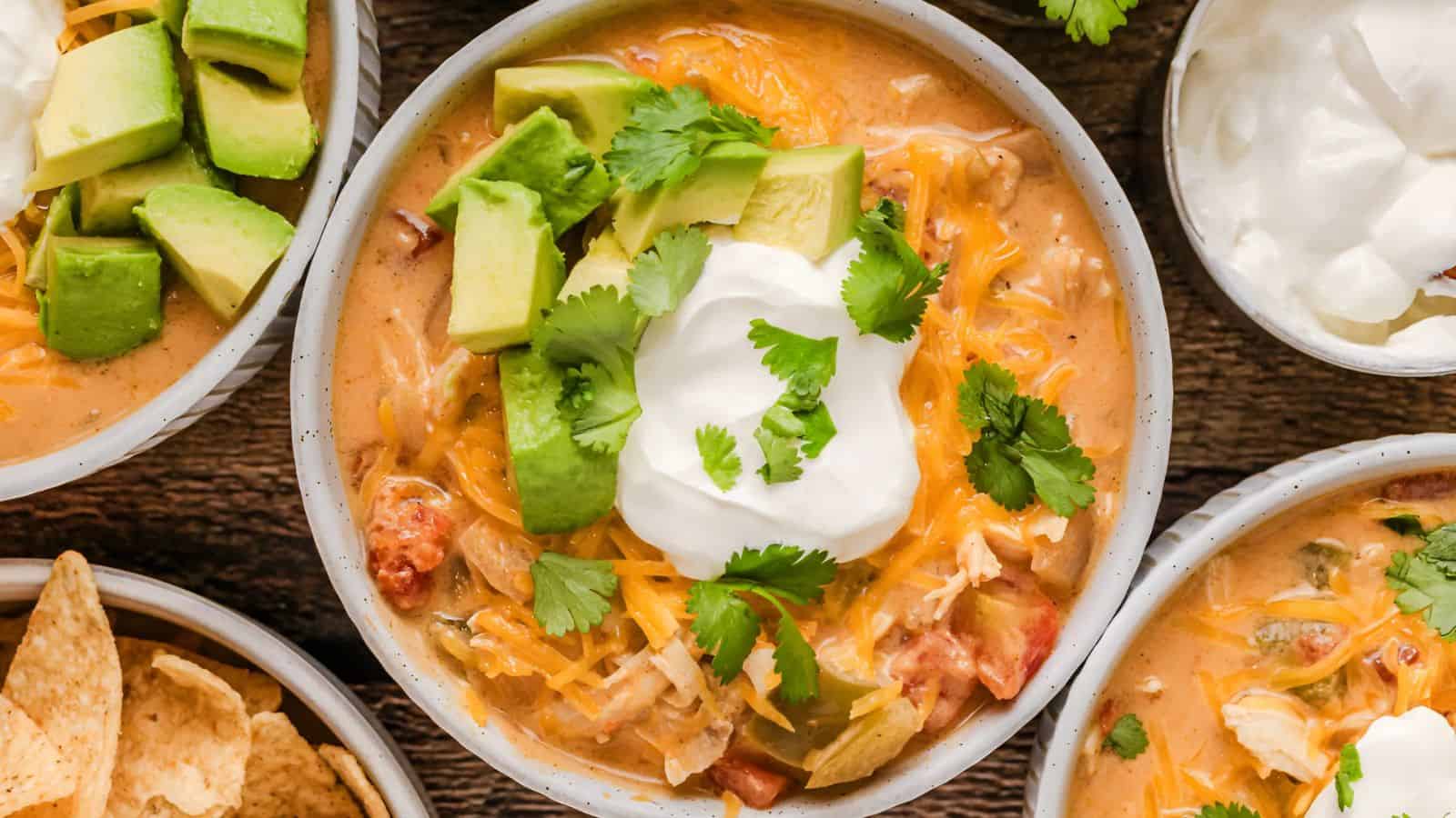 A bowl of chicken tortilla soup topped with avocado, cheese, sour cream, and cilantro, surrounded by bowls of sour cream and chips.