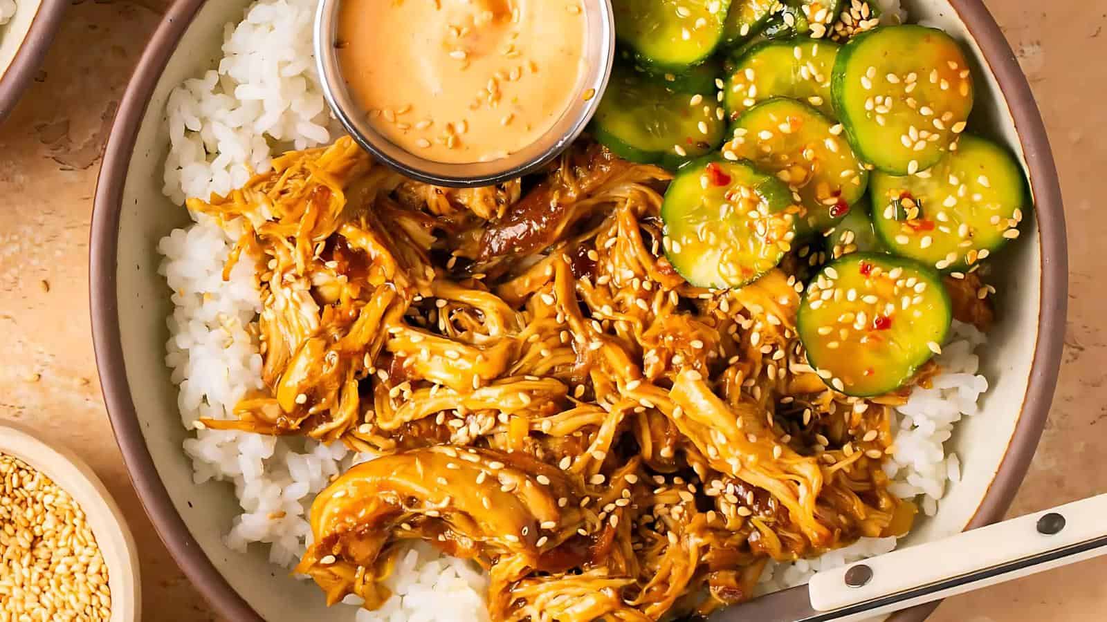 A bowl of white rice topped with shredded chicken in sauce, sliced cucumbers with sesame seeds, and a small cup of dipping sauce.