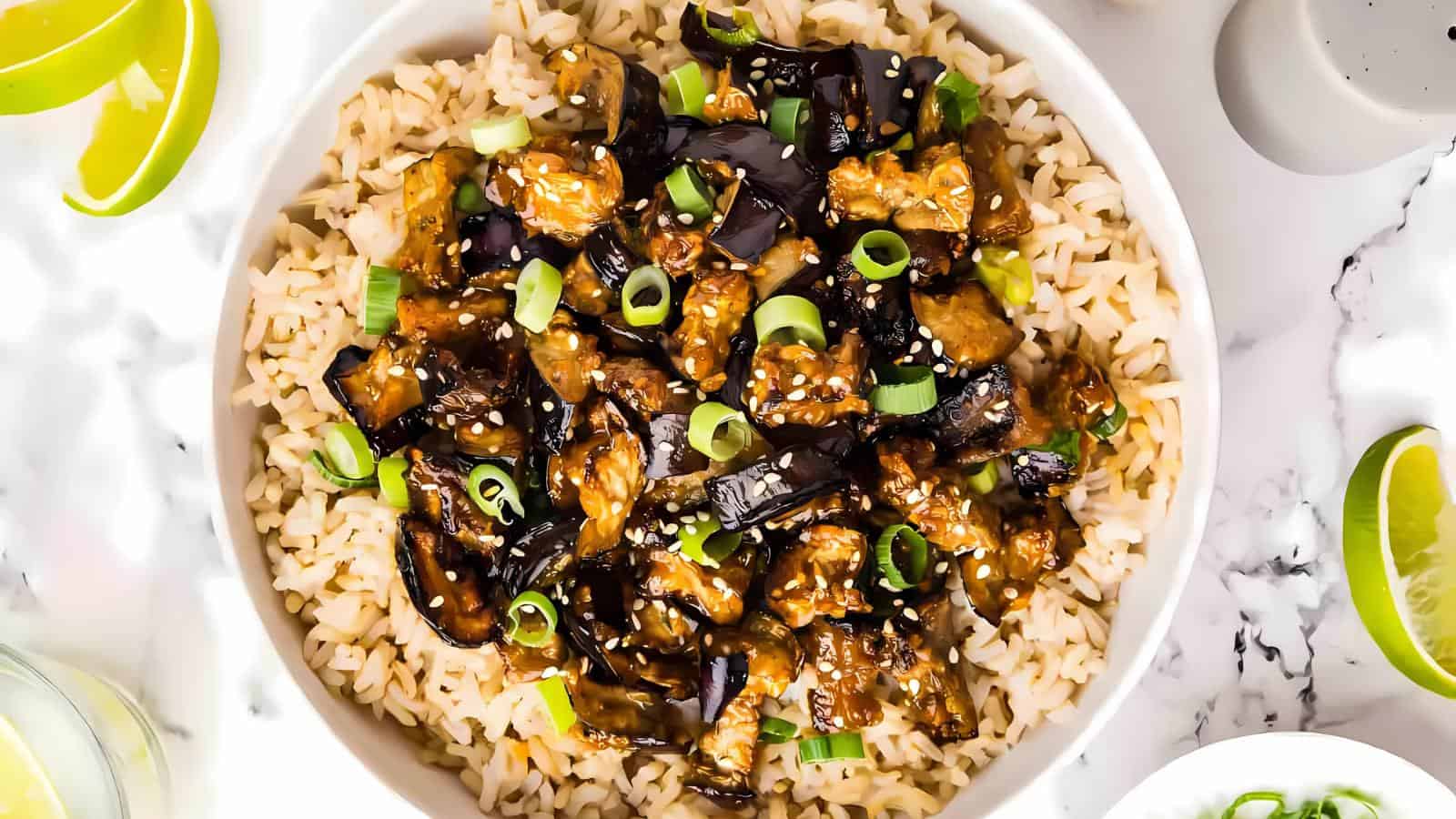 A bowl of rice topped with glazed eggplant, garnished with sliced green onions and sesame seeds. Lime wedges are placed around the bowl on a marble surface.