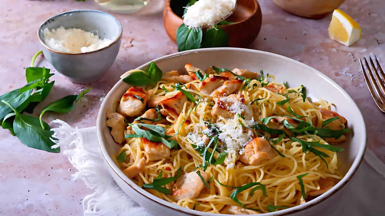 A bowl of spaghetti with grilled chicken, topped with grated cheese and fresh basil. Sides include a small bowl of grated cheese and a wedge of lemon.
