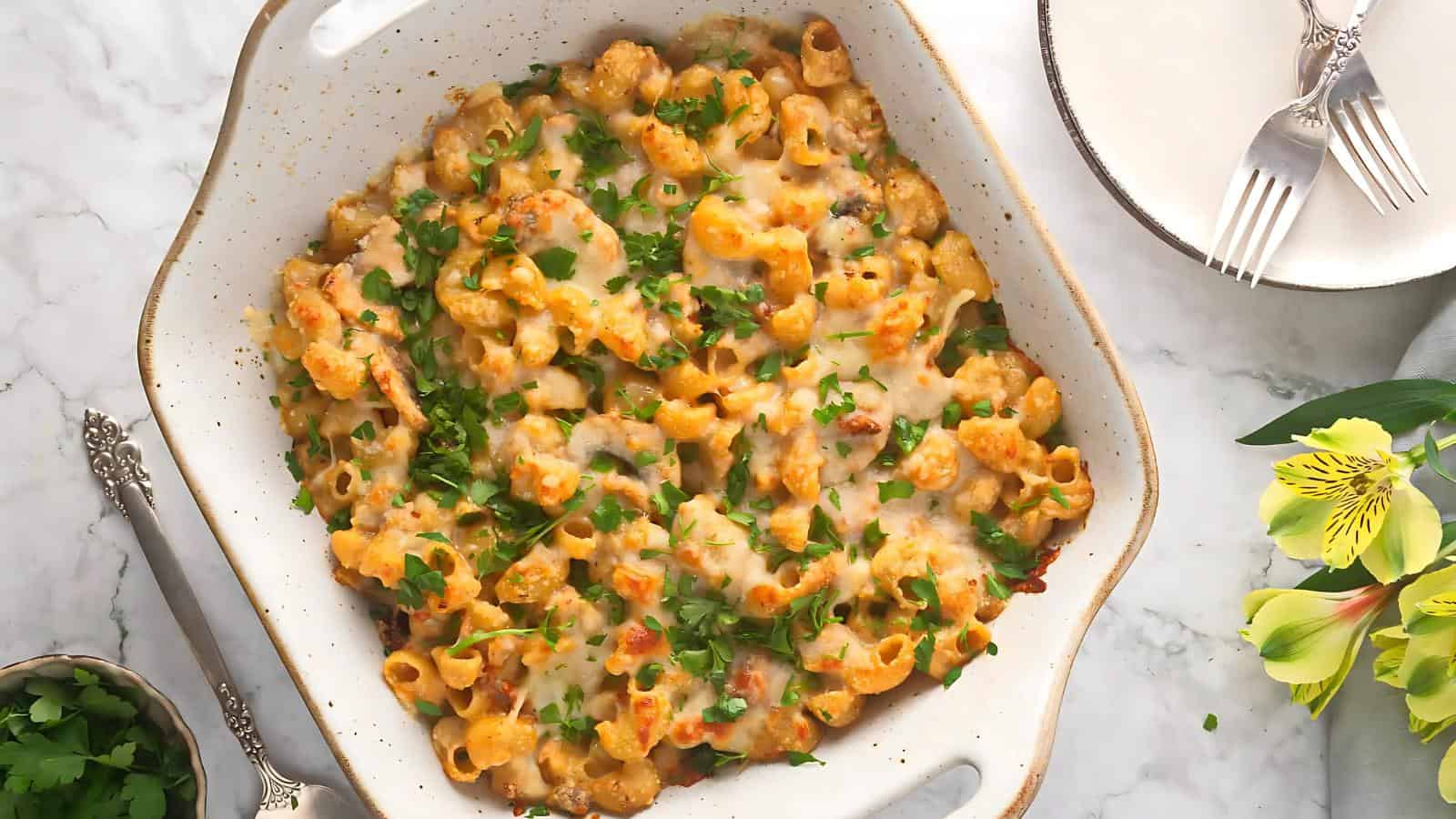 Baked pasta with melted cheese and herbs in a rectangular dish, next to a plate, utensils, and yellow flowers on a marble surface.