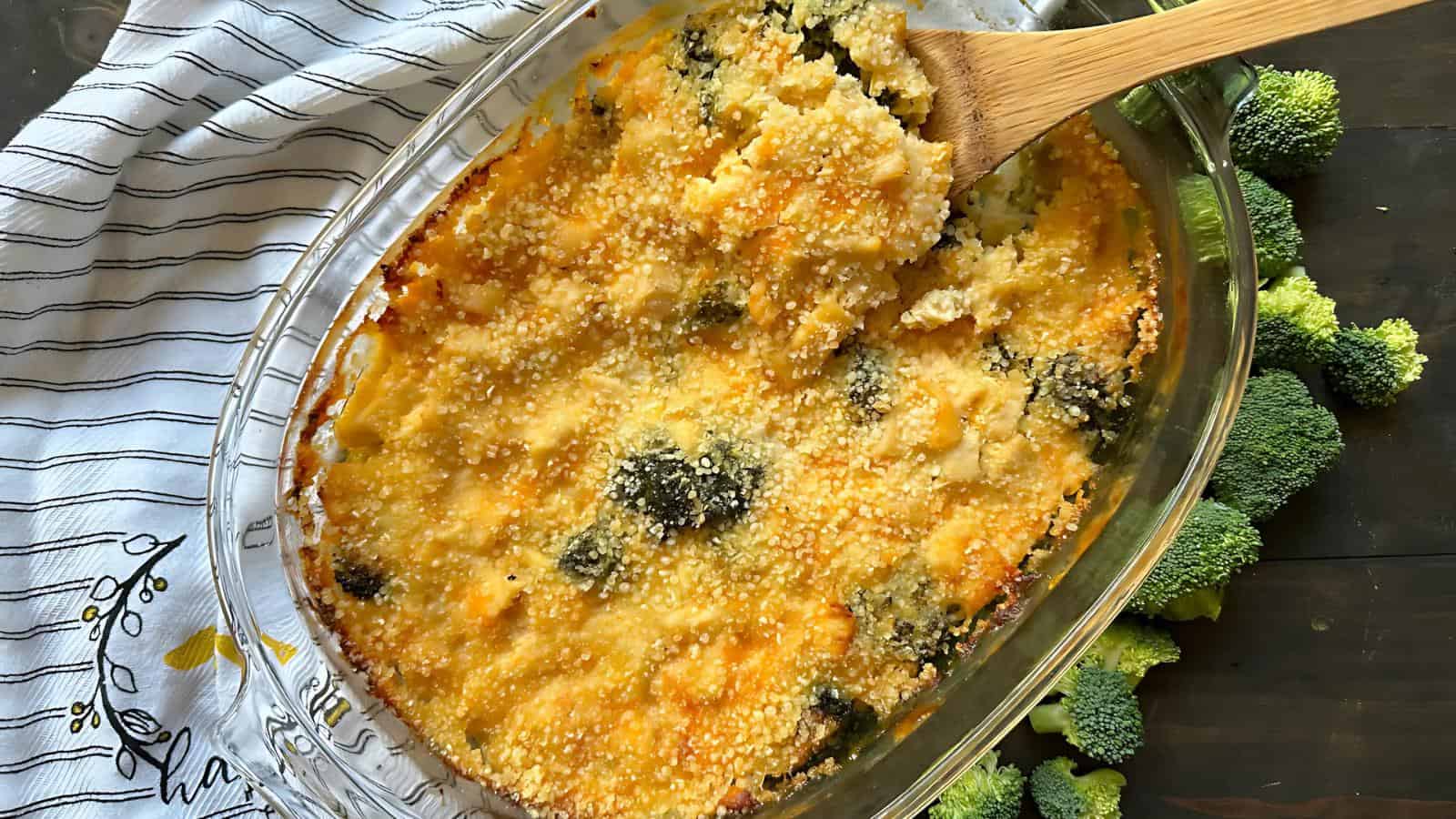 A broccoli and cheese casserole in a glass dish with a wooden spoon, surrounded by fresh broccoli on a dark surface.