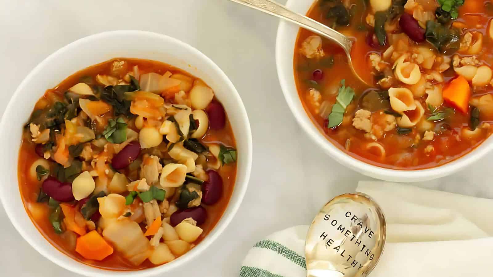 Two bowls of vegetable and pasta soup with beans and greens on a white surface, accompanied by a spoon and a striped napkin.