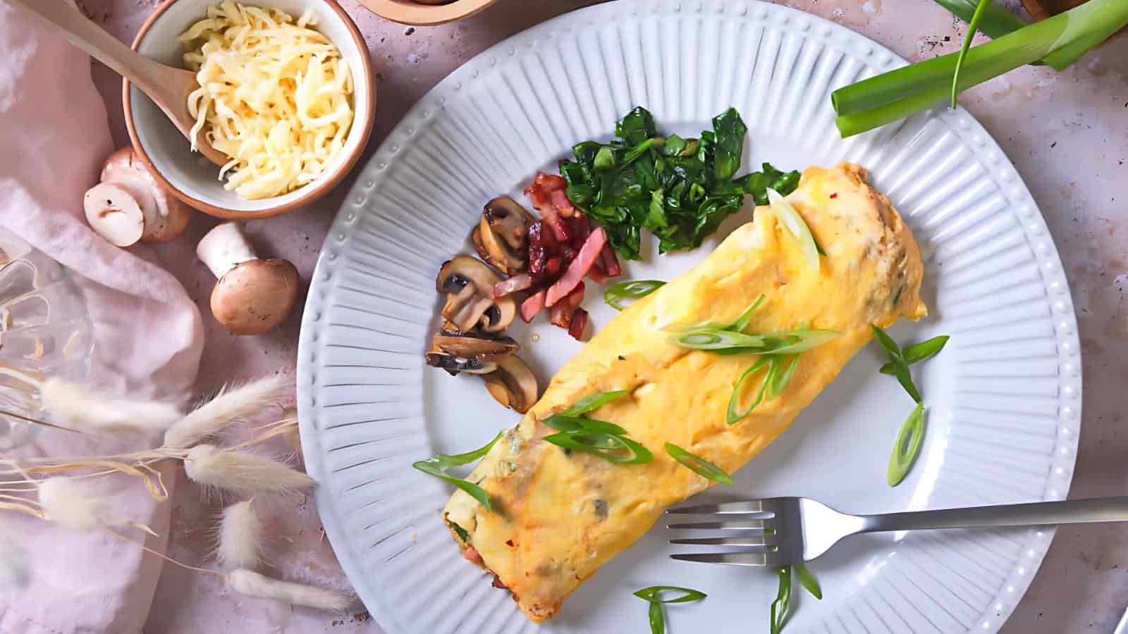 A stuffed omelette on a white plate, garnished with green onions, alongside sautéed mushrooms, greens, and bacon bits. A fork is placed on the plate. Shredded cheese and mushrooms are beside it.