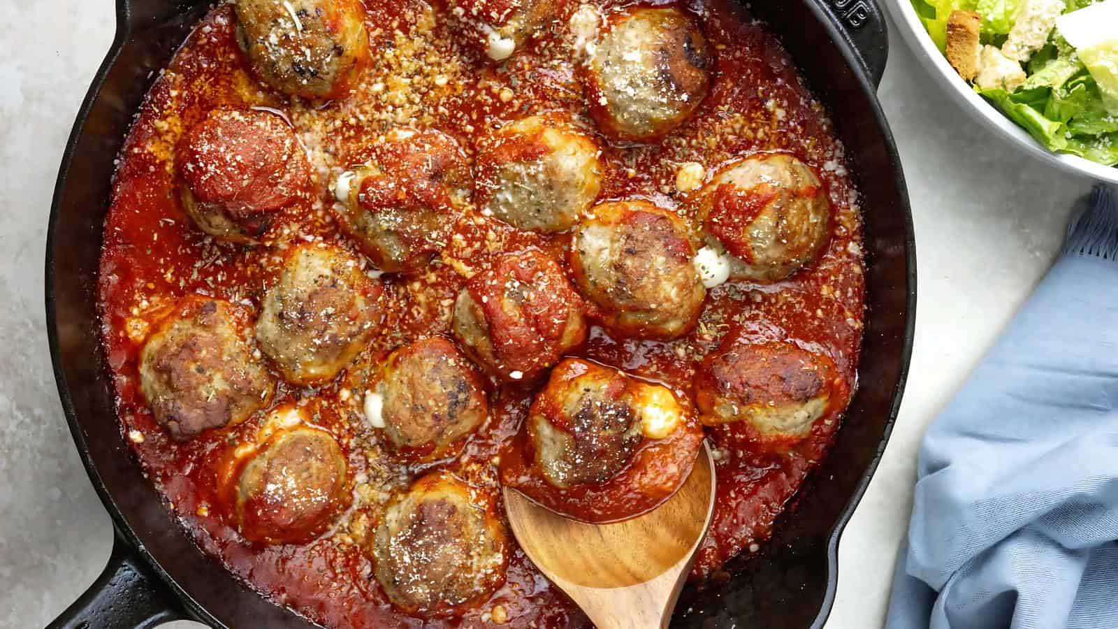 Meatballs in tomato sauce in a skillet with a wooden spoon, next to a blue napkin and a bowl of salad.