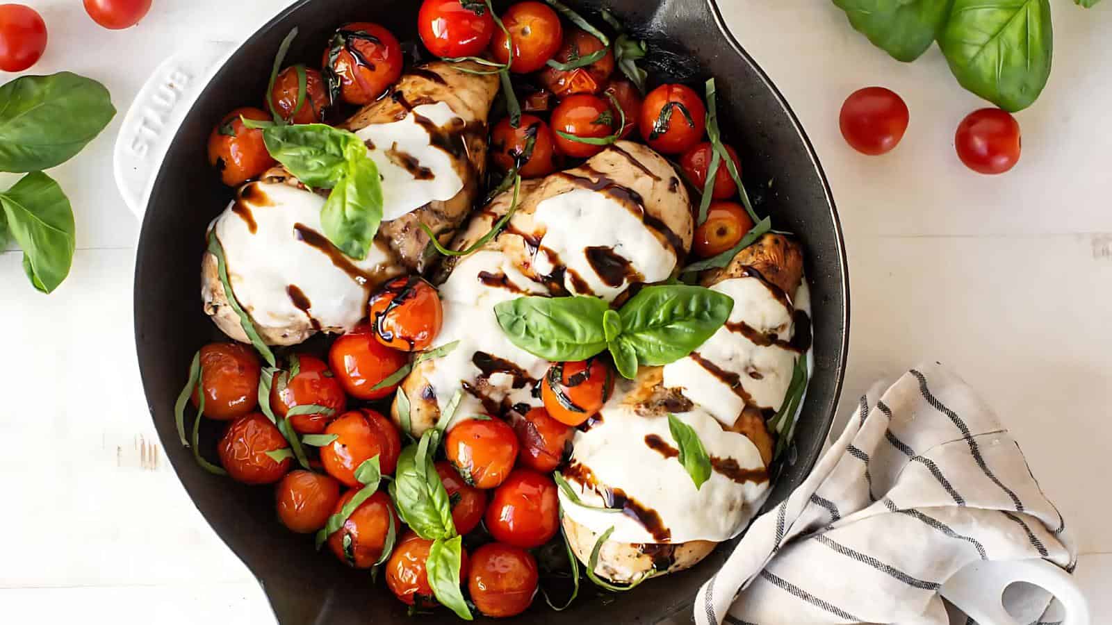 Skillet with grilled chicken breasts topped with melted cheese and balsamic glaze, surrounded by cherry tomatoes and fresh basil leaves. A striped cloth is beside the skillet.