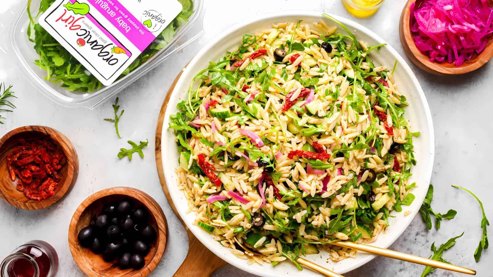 A bowl of orzo salad with arugula, sundried tomatoes, red onions, and olives, surrounded by small bowls of ingredients and a container labeled "organic garden salad.