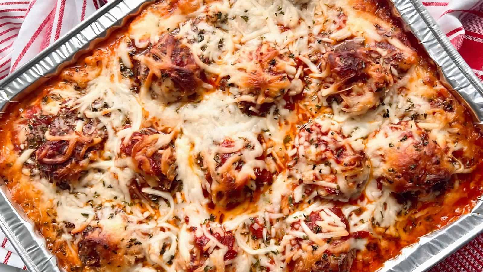 A tray of baked meatball parmesan topped with melted cheese and herbs in a foil container.