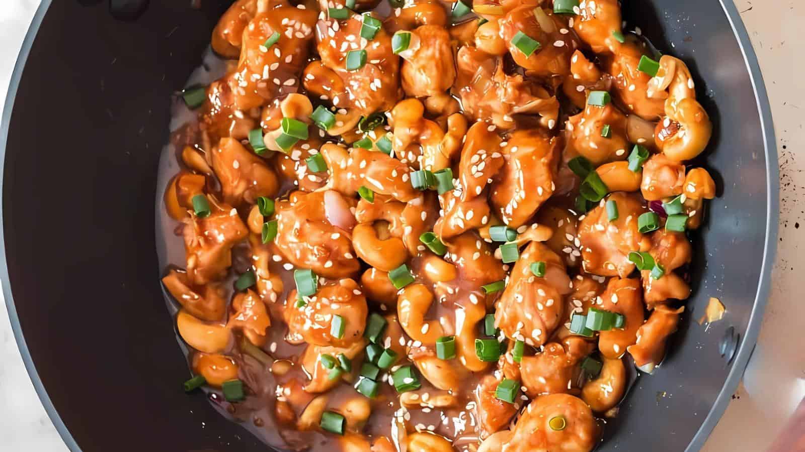 Stir-fried cashew chicken in a black pan, topped with sesame seeds and green onions.