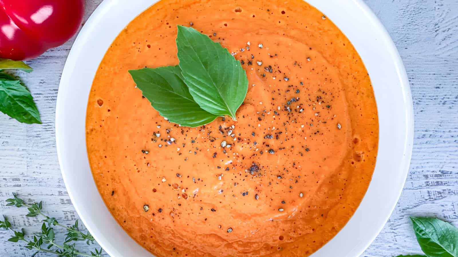 A bowl of creamy tomato soup garnished with fresh basil leaves and black pepper, surrounded by a red bell pepper and herbs on a light wooden surface.