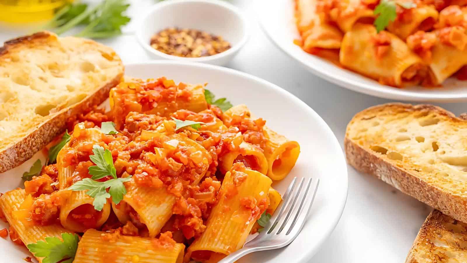 A plate of rigatoni with tomato sauce, garnished with parsley, is served with toasted bread. A small bowl of chili flakes is visible in the background.