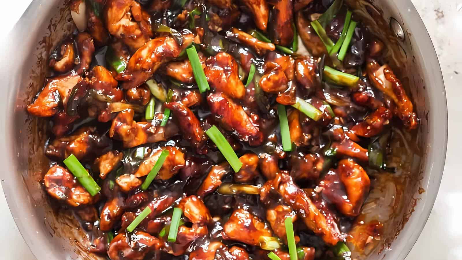 A close-up of a dish with glazed chicken pieces and green onions in a thick, dark sauce.