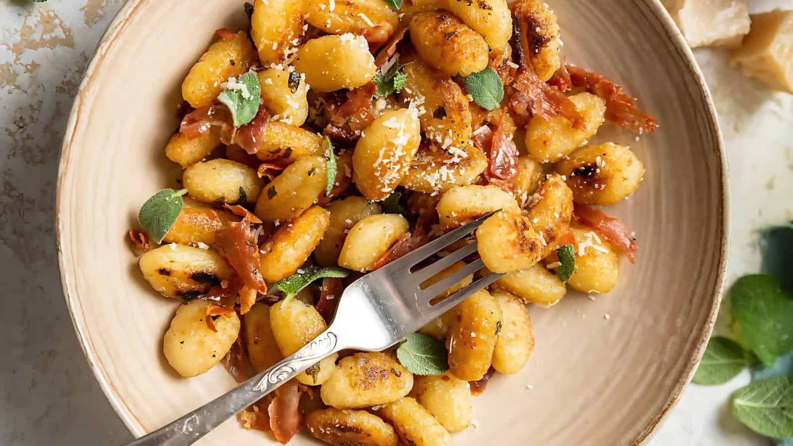 A plate of browned gnocchi with crispy prosciutto, garnished with fresh sage and grated cheese. A fork rests on the side.