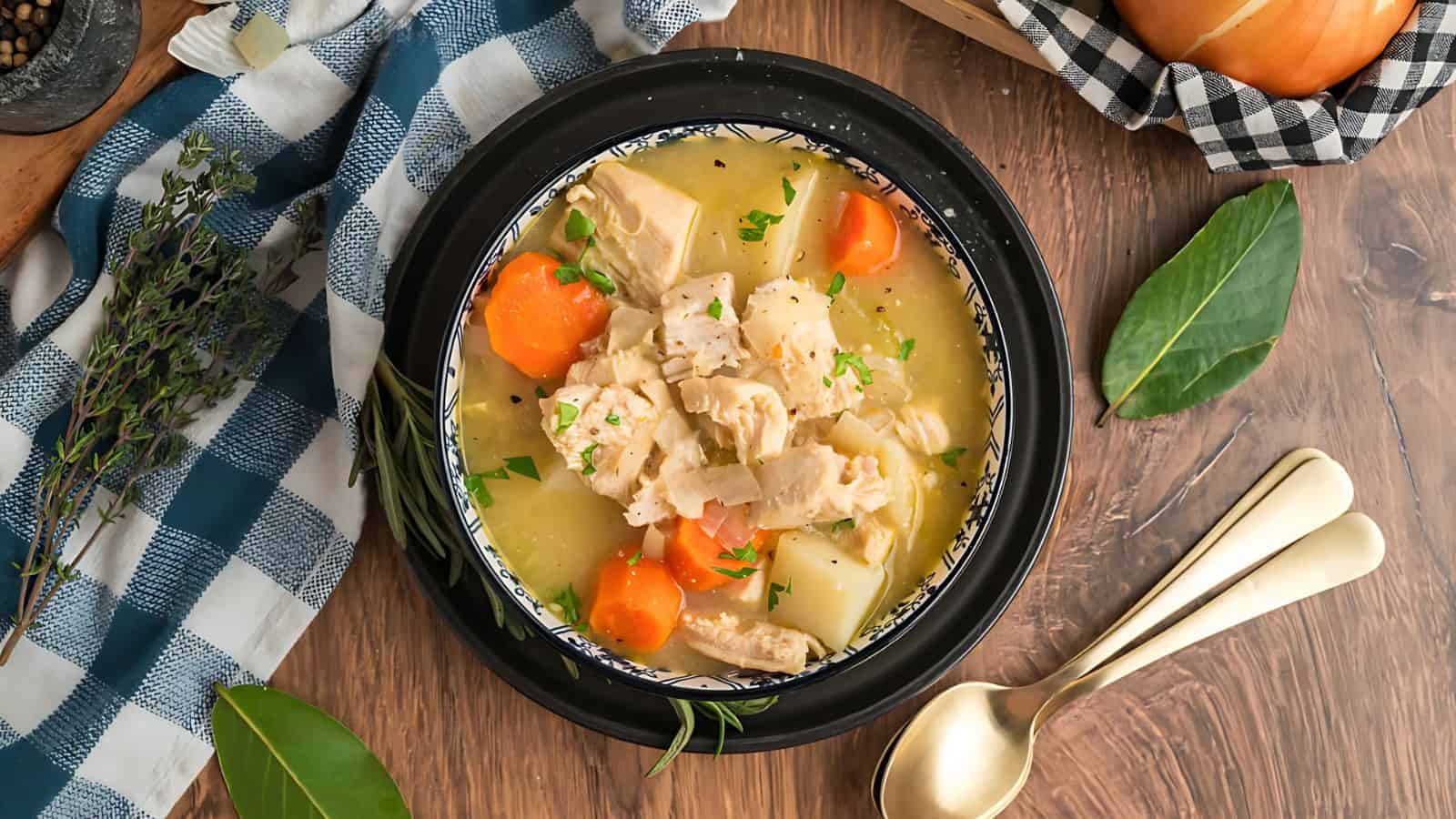 A bowl of soup with chunks of chicken, carrots, and potatoes on a wooden table. A checked cloth napkin and two spoons are beside the bowl.
